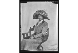 Woman wearing a skirt, jacket, and hat sitting in a chair. Image is black and white.